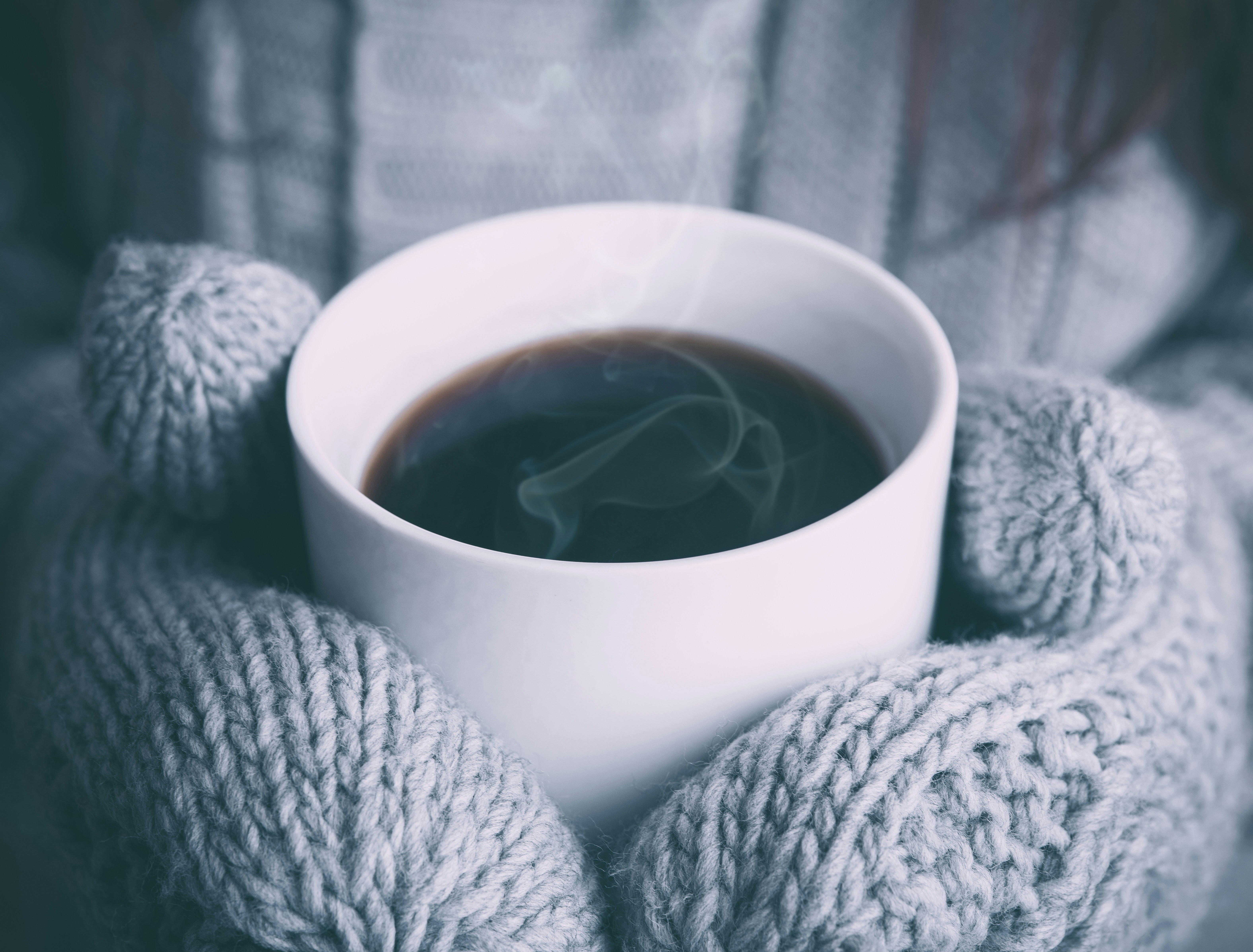 Close-up of a pair of hands wearing thick, gray knitted gloves holding a white ceramic cup filled with steaming coffee. The background is blurred, with a matching gray knitted sweater visible, creating a cozy and warm atmosphere. The steam rising from the coffee adds a sense of warmth and comfort, evoking feelings of relaxation and serenity on a cold day.