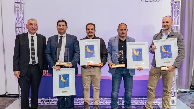 Five men pose during an award ceremony, standing in front of a backdrop with IST logo and Spanish tagline "Tu mutualidad!" Three recipients hold framed certificates featuring a crescent moon design and one also holds a trophy. The men are dressed in formal and business casual attire. The event celebrates achievements in a professional setting.