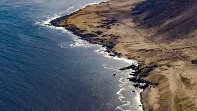 An aerial view of the coastline near Taltal, Chile. The image features a dramatic meeting of the Pacific Ocean and arid desert terrain, with rugged rocky shores and sandy hills extending into the distance. A winding dirt road follows the edge of the coast, with waves gently lapping against the shore under a hazy sky.