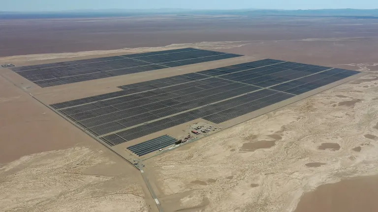 Aerial view of a large solar farm in the Atacama Desert, Chile, featuring expansive rows of dark solar panels covering vast sections of the arid landscape. The barren desert terrain surrounds the facility, with a small operations area visible near the bottom. The distant mountains and a clear blue sky can be seen in the background.