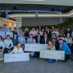 A group of approximately 25 young adults are posing for a photo in an office setting. Some are holding large novelty checks with various amounts written on them. The individuals are diverse in appearance and are dressed in business casual attire.  Artificial greenery lines the wall. The mood of the image is celebratory and suggests a competition or award ceremony.