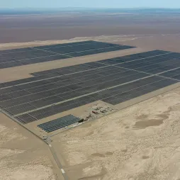 Aerial view of a large solar farm in the Atacama Desert, Chile, featuring expansive rows of dark solar panels covering vast sections of the arid landscape. The barren desert terrain surrounds the facility, with a small operations area visible near the bottom. The distant mountains and a clear blue sky can be seen in the background.